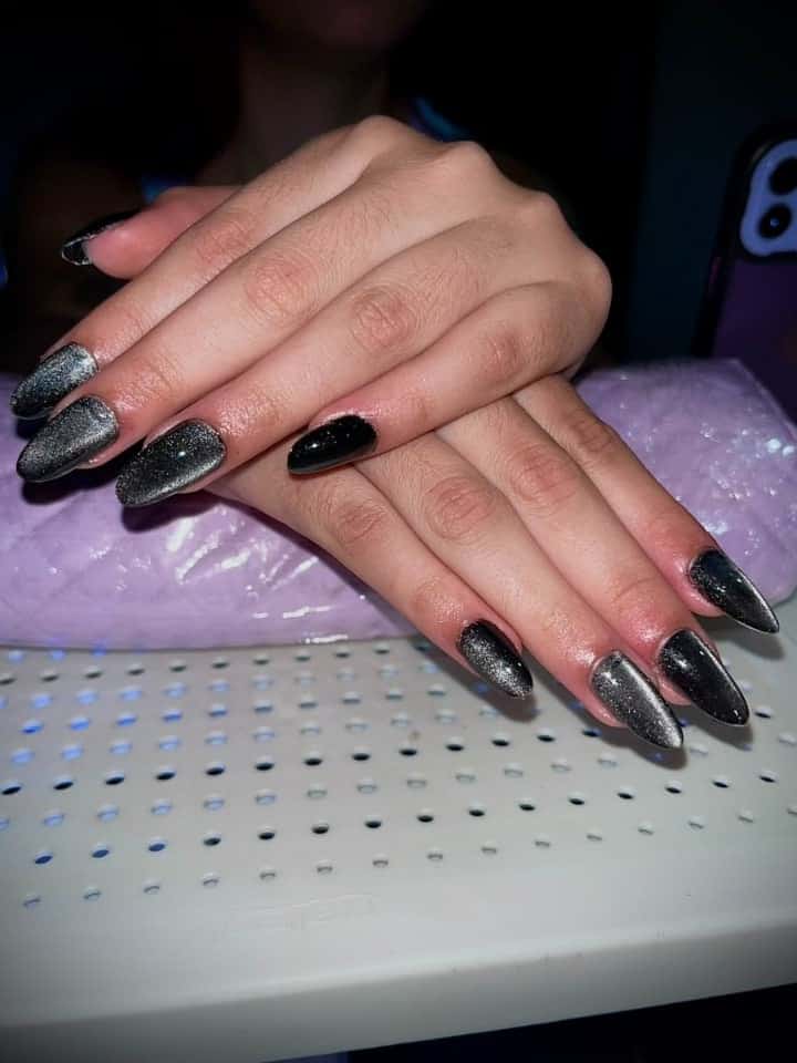 Duas mãos cruzadas com unhas amêndoa longas em gel magnético preto, com risco prateado atravessando cada unha